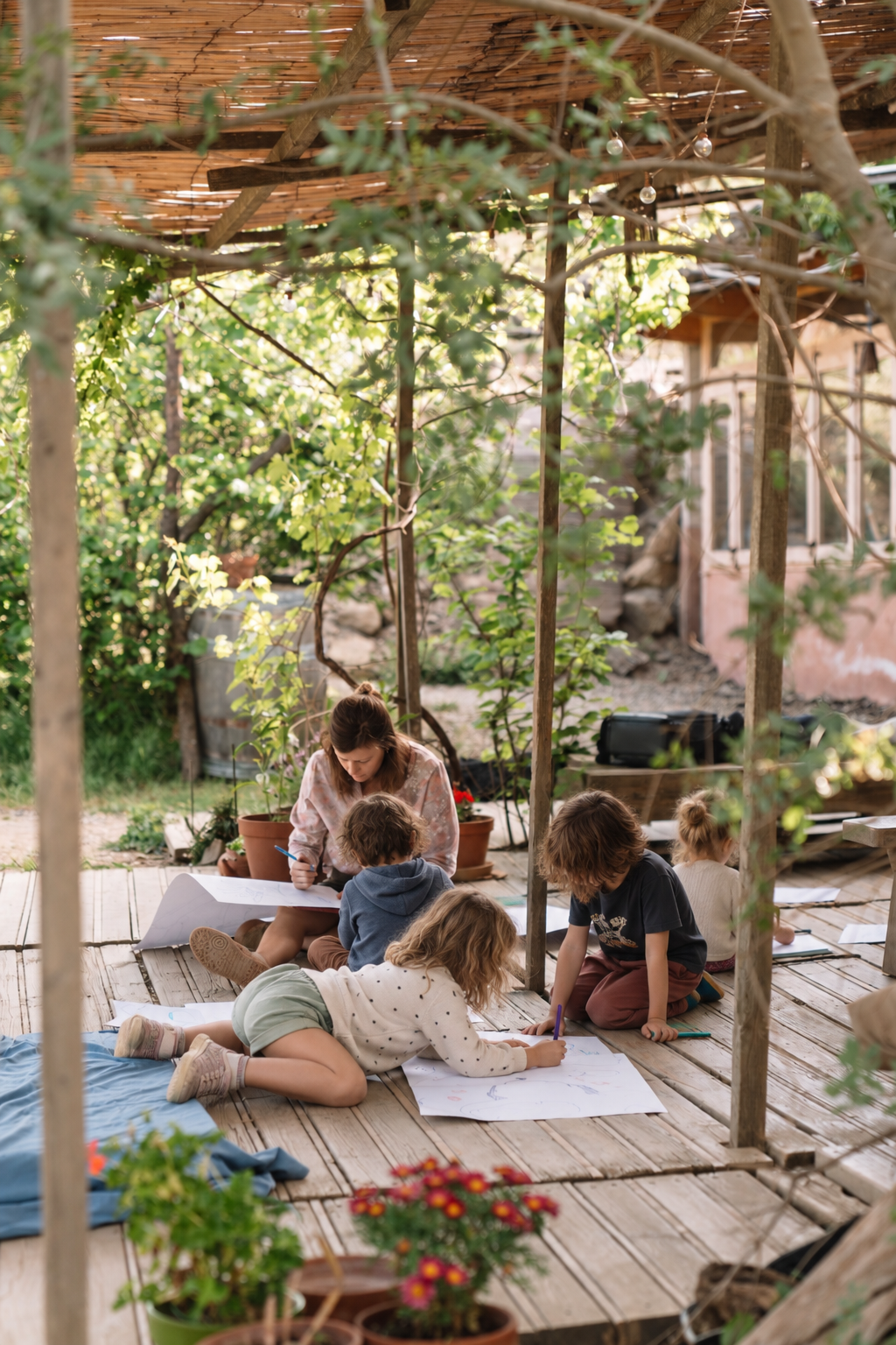 Camille drawing with children on a wooden deck under a vine-covered pergola surrounded by greenery